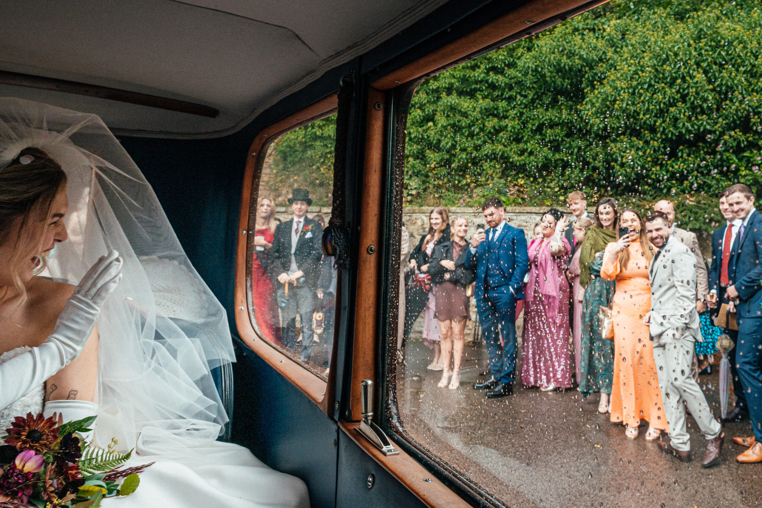 A bride waves from inside a vintage car as guests in colourful outfits watch - by UK documentary wedding photographer Simon Dewey