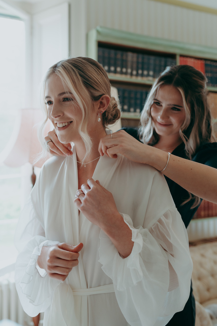 A bride getting ready has a necklace put on for her by a bridesmaid. They're both smiling as they look into a mirror. - by Damien Vickers Photography