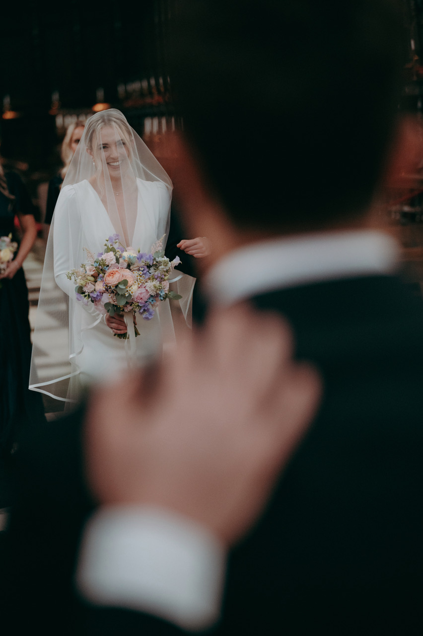 A bride smiles as she approaches her groom carrying a bouquet of colourful pastel flowers. In the foreground and beautifully blurred, the groom faces her, with a friend's hand on his shoulder - by Damien Vickers Photography in Cambridge