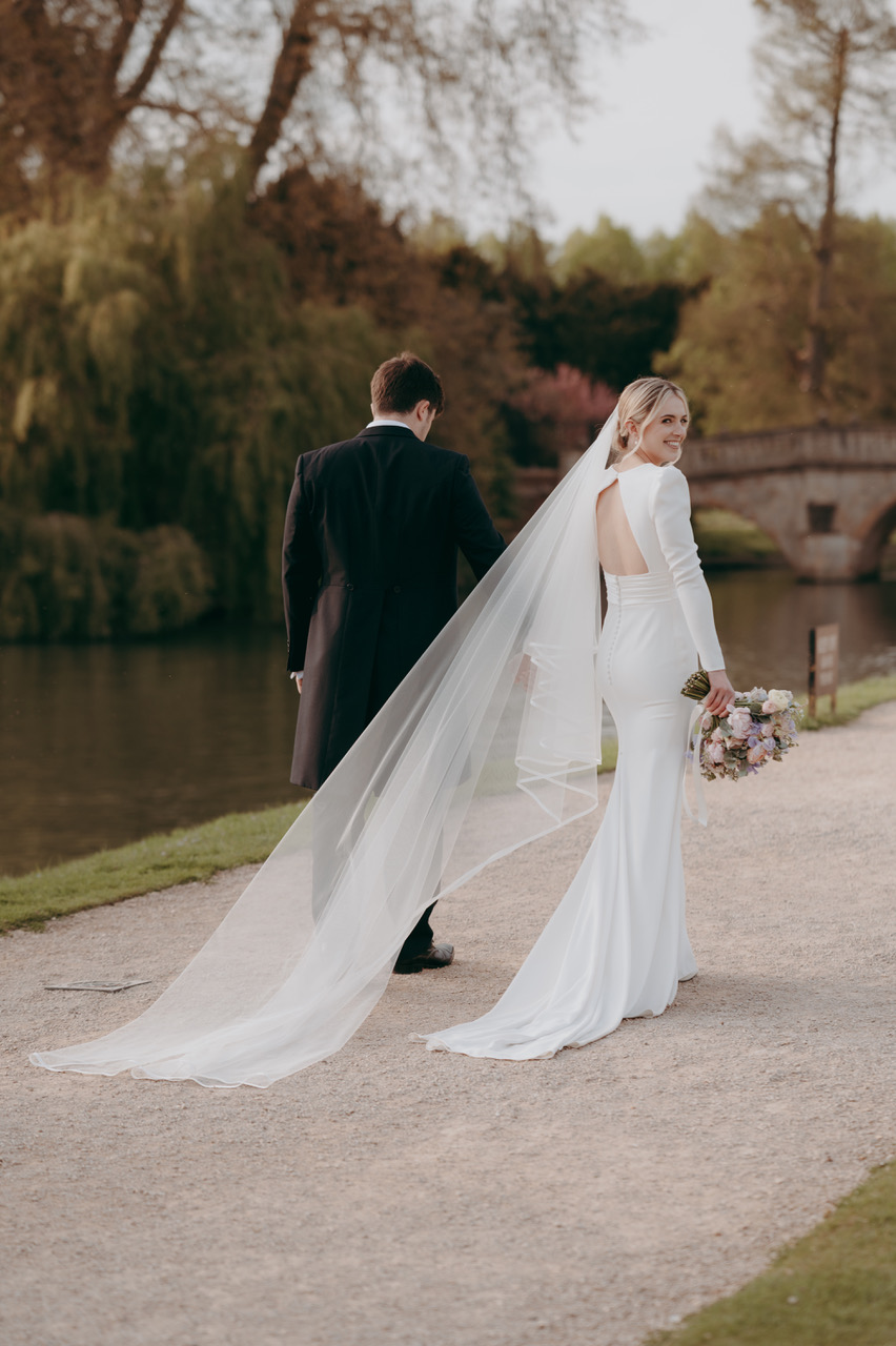 A groom and bride walk away from the camera along the riverside path in Cambridge. The bride turns slightly towards the camera. She's wearing a backless dress with long sleeves and her veil trails behind her. - by Damien Vickers Photography in Cambridge