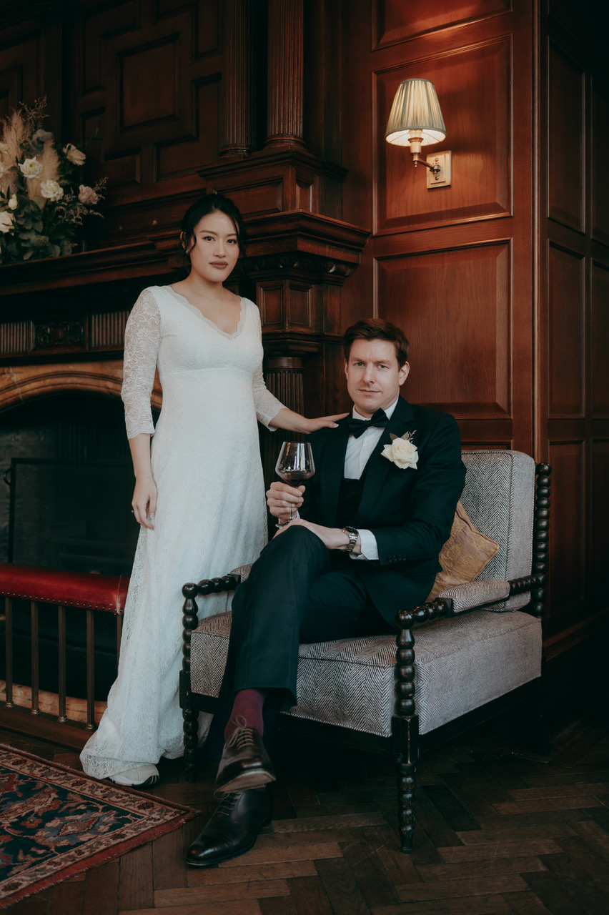 Portrait of a bride and groom in a wood-panelled stately home. He's sat on a chair while she stands to his left with a hand on his shoulder. - by Damien Vickers Photography