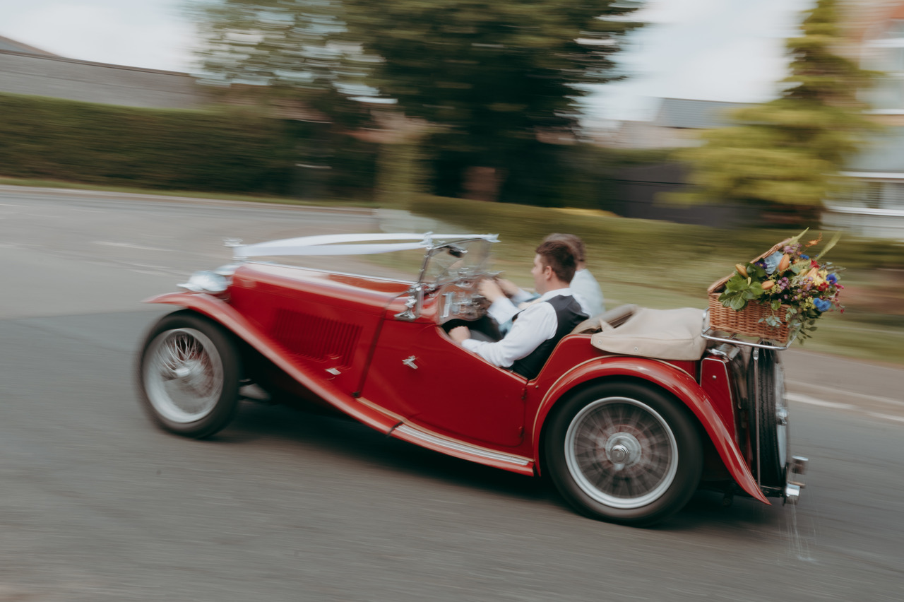Two men in a moving vintage sports car with a basket of flowers on the back and a white ribbon on the front - by Damien Vickers Photography