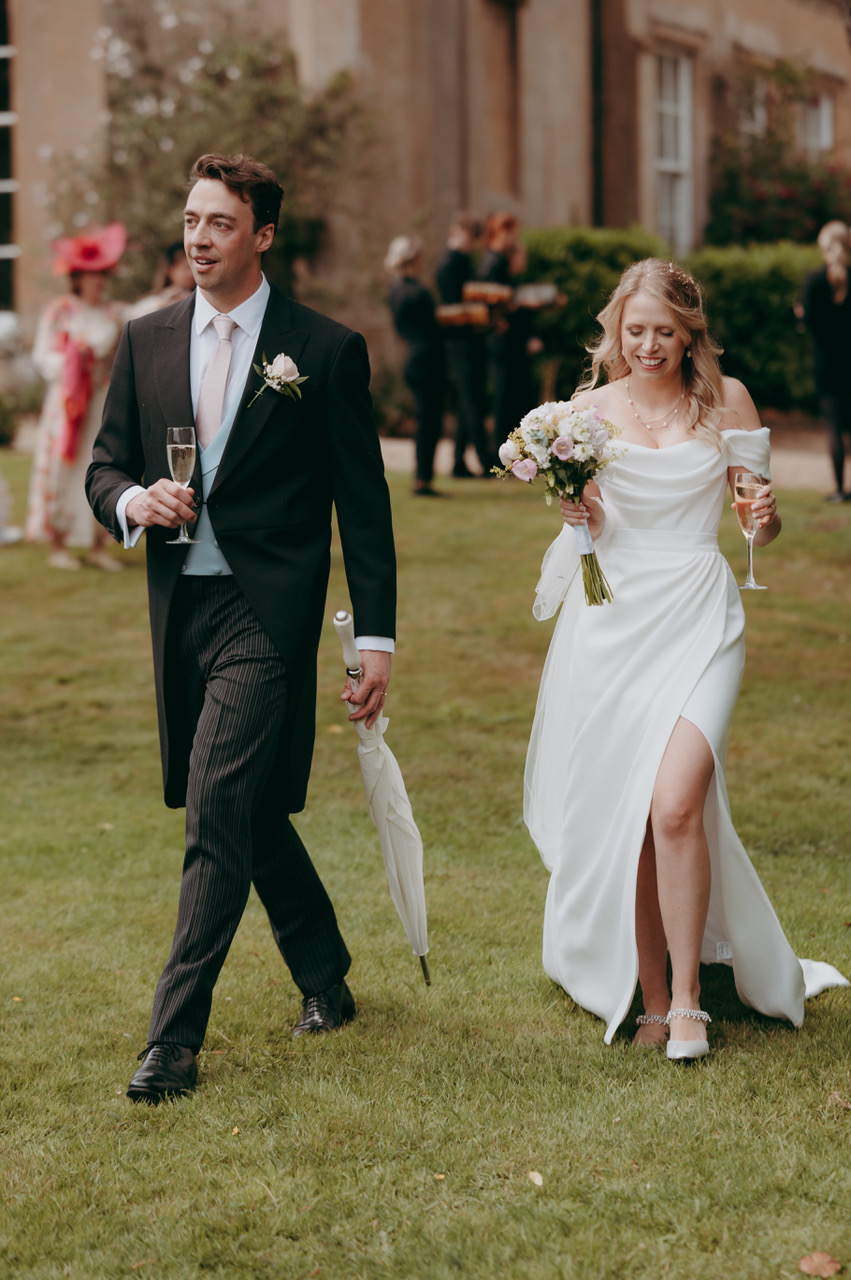 A groom and bride walk across the grass outside their venue. Both are holding champagne flutes, he's also got a folded white umbrella and she carries a bouquet - by Damien Vickers Photography in Cambridge