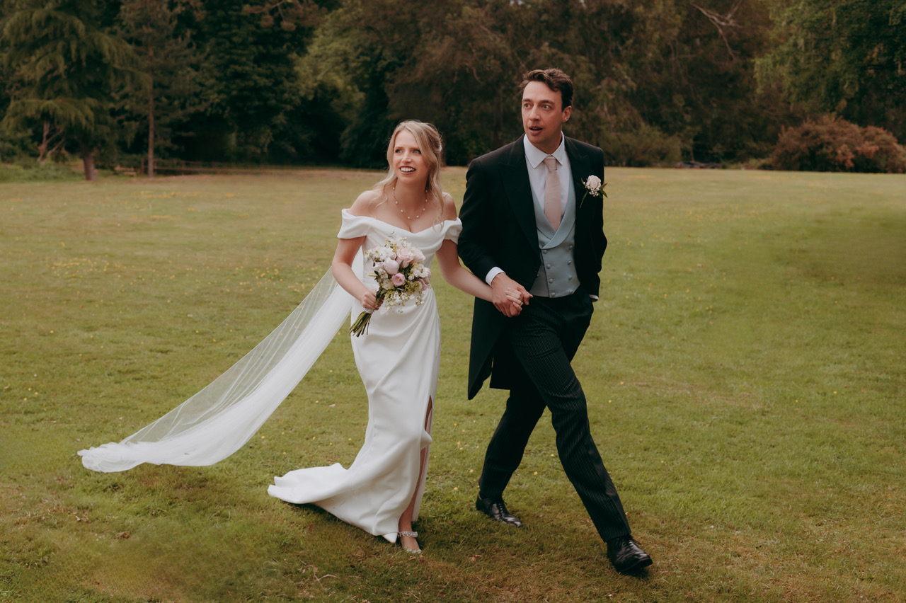A bride and groom walking across a grassy lawn and looking at something to the left of the camera. She's holding a petite bouquet of pale pink flowers - by Damien Vickers Photography