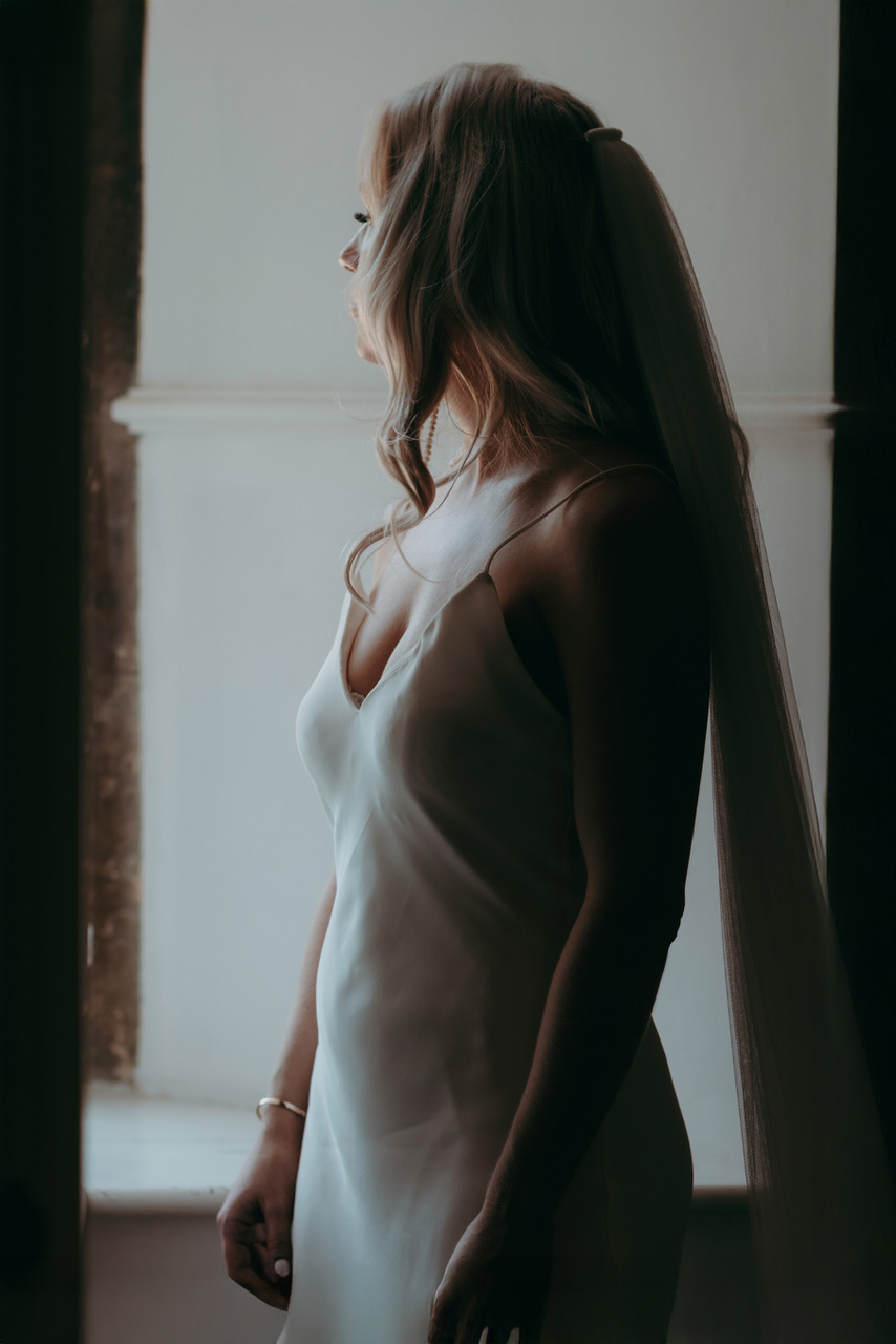 A bride stands by a window with soft natural light highlighting her features - by Damien Vickers Photography in Cambridge