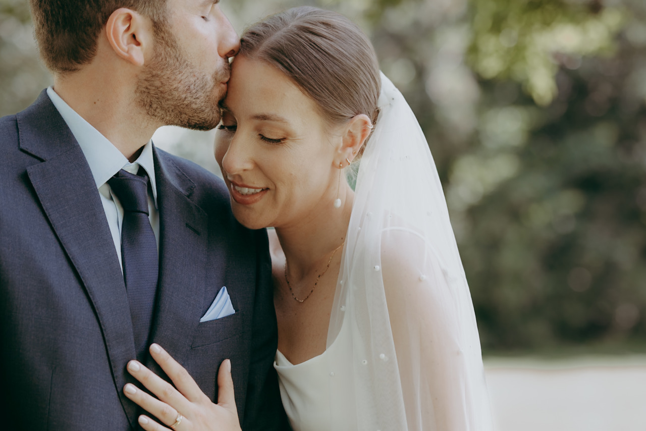 A natural moment between groom and bride as he kisses her forehead. She rests a hand on his chest. He's wearing a navy suit and she's in a white stripy dress with embroidered veil - by Damien Vickers Photography in Cambridge