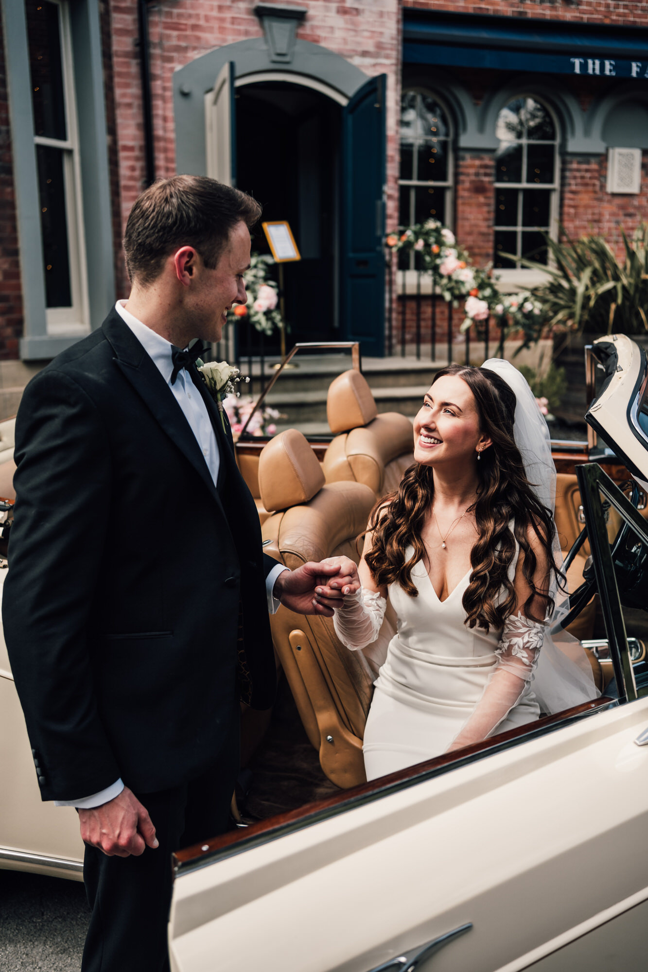 A groom and bride smile at each other as he holds her hand to help her from a vintage wedding car. Natural wedding portraits in Yorkshire by David Lindsley Photography