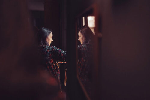 A woman looking thoughtfully out of a window, her image reflected in the glass. Claire Gould by Tiree Dawson Photography