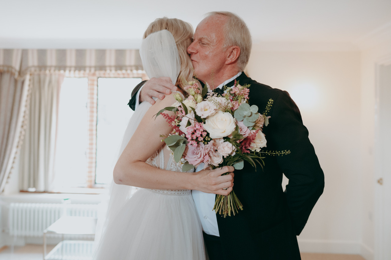 A proud dad kisses his daughter on the cheek before her wedding. She's holding a bouquet of ivory and blush roses and spring flowers - by Damien Vickers Photography in Cambridge