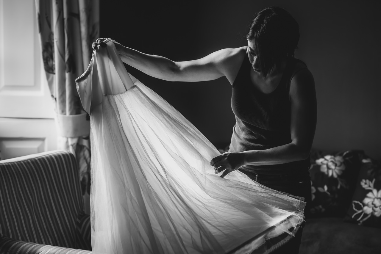 A moment of stillness before a wedding, as a woman lifts a sheer dress from an armchair, catching the natural light from a nearby window - by Damien Vickers Photography in Cambridge
