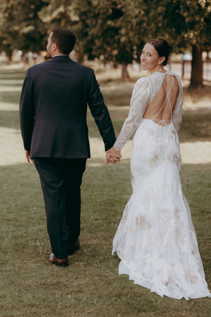 A groom and bride walk away from the camera holding hands. The bride turns towards the camera. She's wearing a delicately embroidered white dress in a backless style with crossover spaghetti straps - by Damien Vickers Photography
