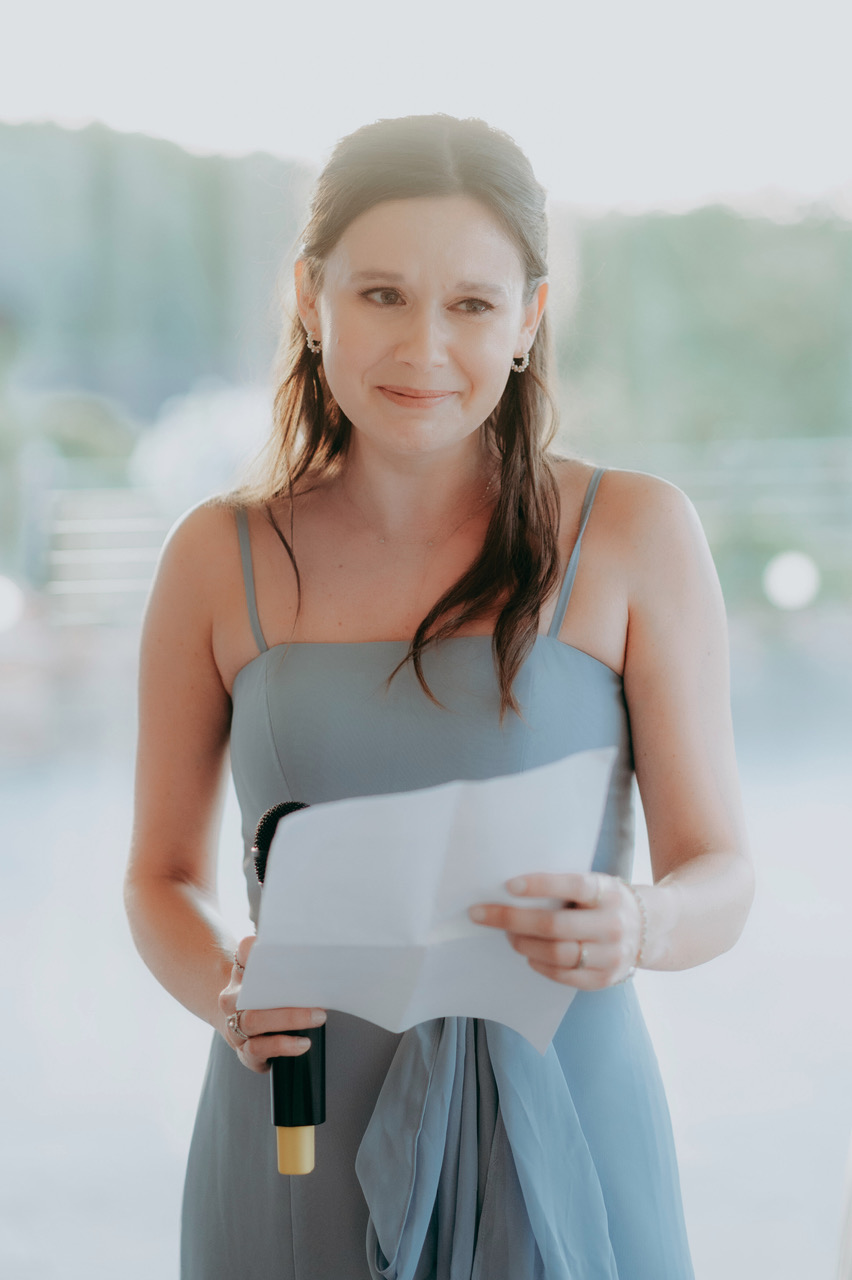 A tearful bridesmaid looks out towards guests as she gives a wedding speech. She's holding a microphone and an unfolded sheet of paper. Behind her, a clear marquee window and the greenery of the countryside - by Damien Vickers Photography in Cambridge