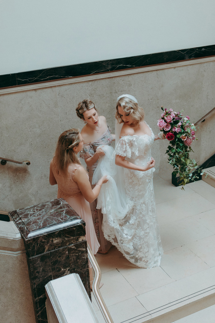 A bride and her maids of honour pause on an elegant staircase. The train of the dress is held up and away from the floor. The bride is holding a bouquet of pink and red roses with trailing greenery - by Damien Vickers Photography in Cambridge
