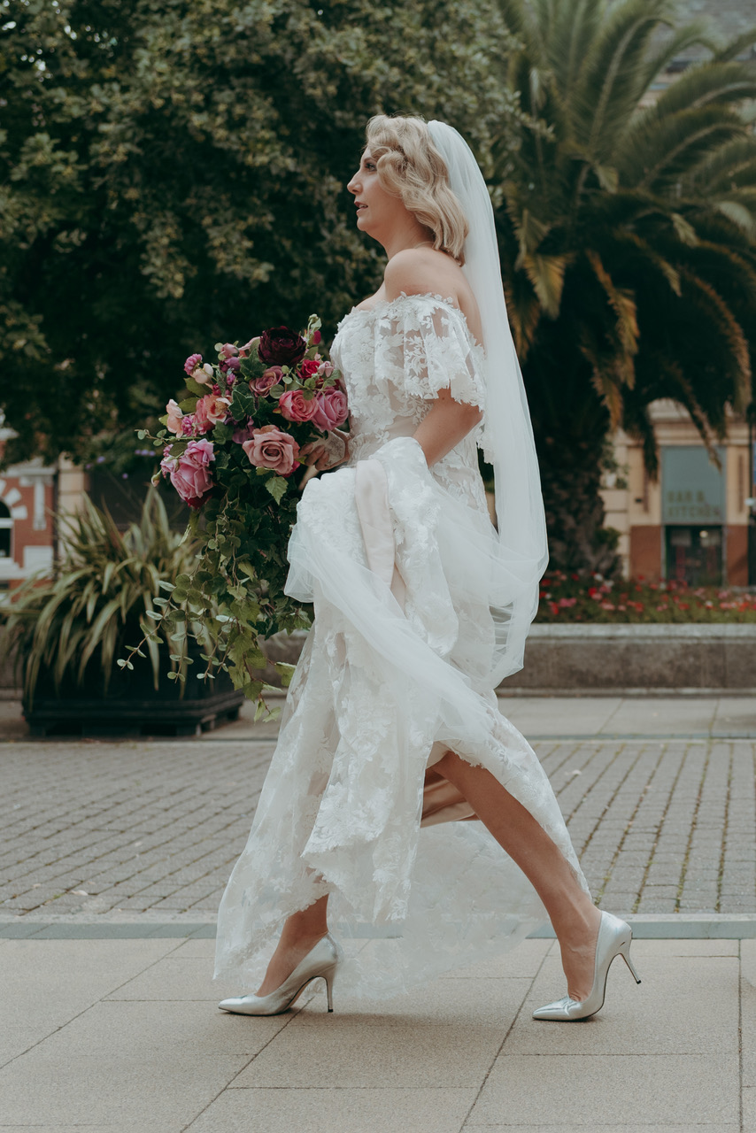A bride walks quickly from right to left across the frame, with her lace dress gathered in one hand and a bouquet of pink roses in the other - by Damien Vickers Photography