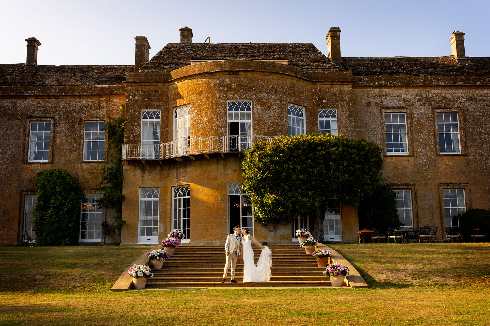 A newlywed couple stand on the steps outside North Cadbury Court at golden hour. The bride leans in for a kiss, holding her dress in the sunlight. By Martin Dabek Photography