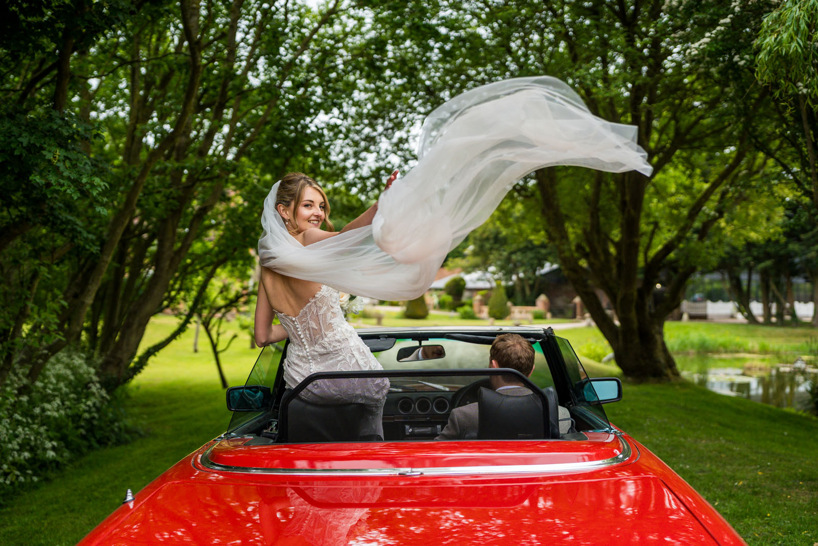 A bride stands in a sports car with the roof open, her veil catching the wind at Solton Manor - Benjamin Toms Photography | Kent