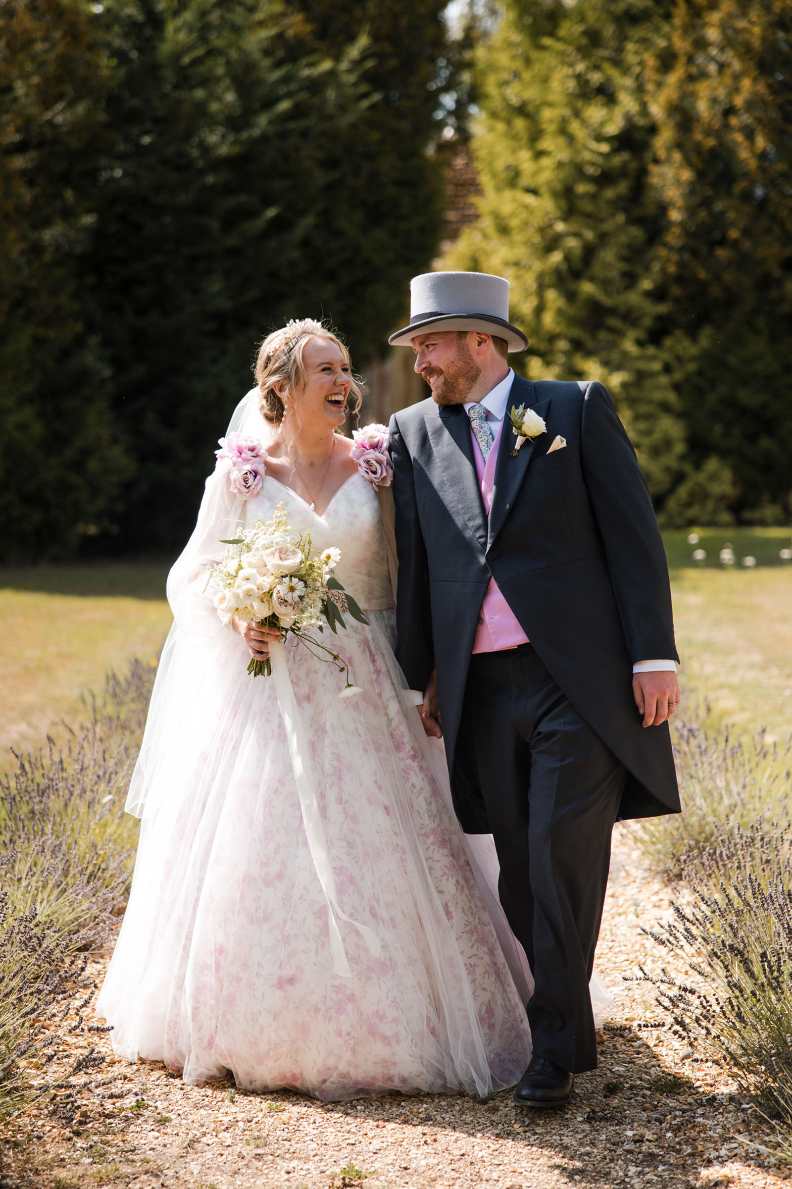 A bride in a floral print dress with life size silk roses on the shoulders, carrying a bouquet of white flowers, hand in hand with a groom in top hat and tails with pink waistcoat - by Wiltshire photographer Greg Shingler Photography