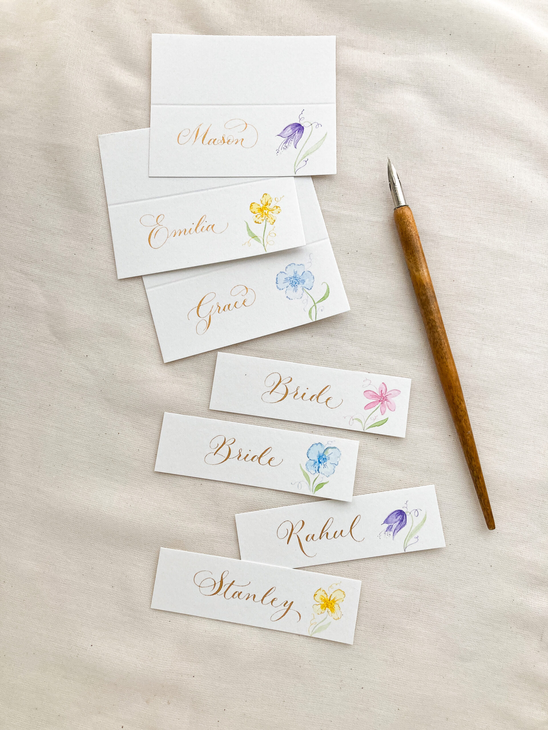 Three folded white wedding place name cards and four elegant slimline wedding place name tags, all with hand written calligraphy and delicate watercolour flower illustrations | By Moon & Tide Calligraphy UK