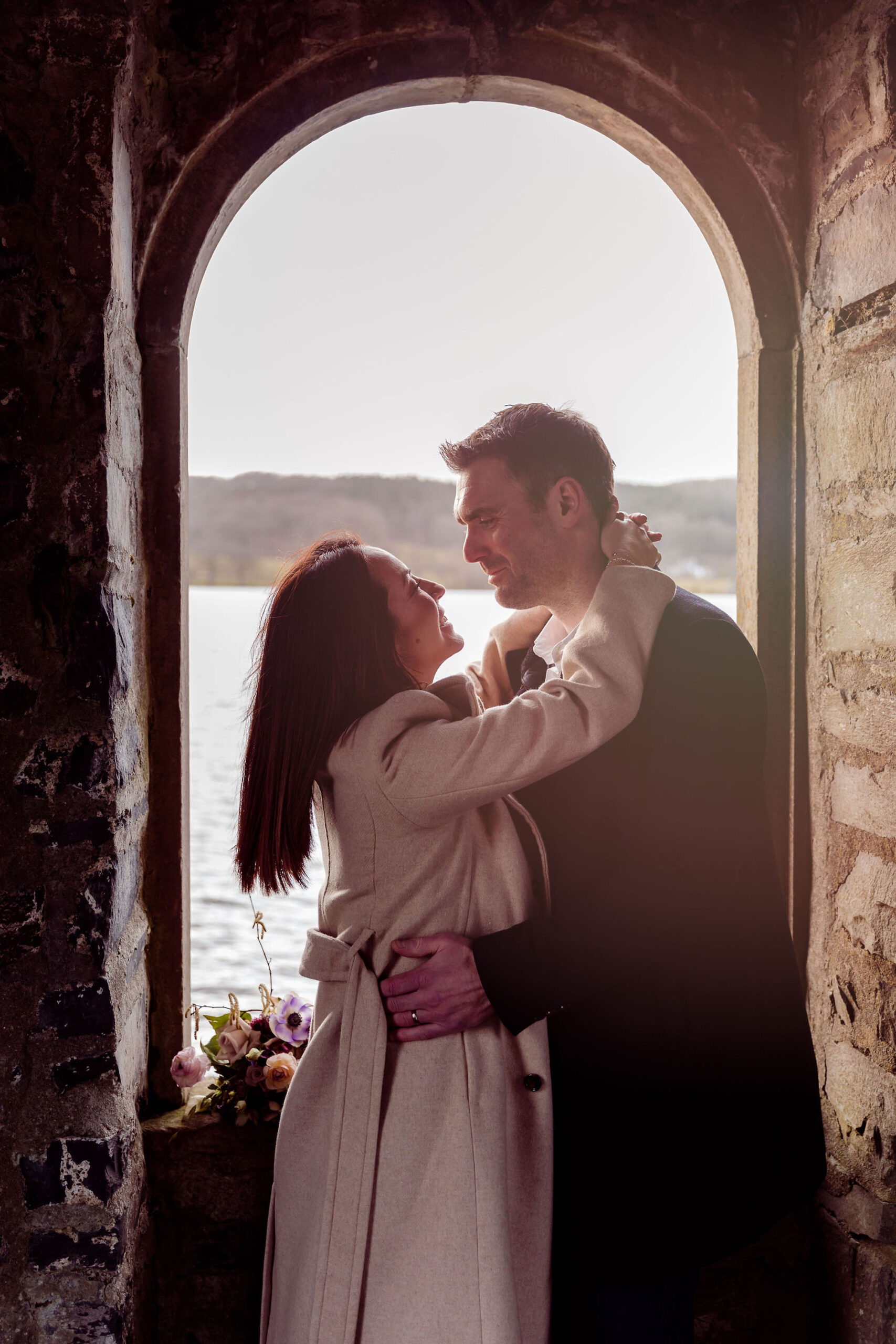 Couple embracing under a stone archway with Lake Windermere and mountains in the background | Mark Battista Photography