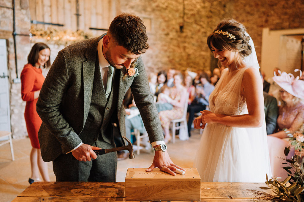 Man with a hammer nailing closed a decorative wooden box during a wedding ceremony. A woman in a wedding dress smiles as she watches him.