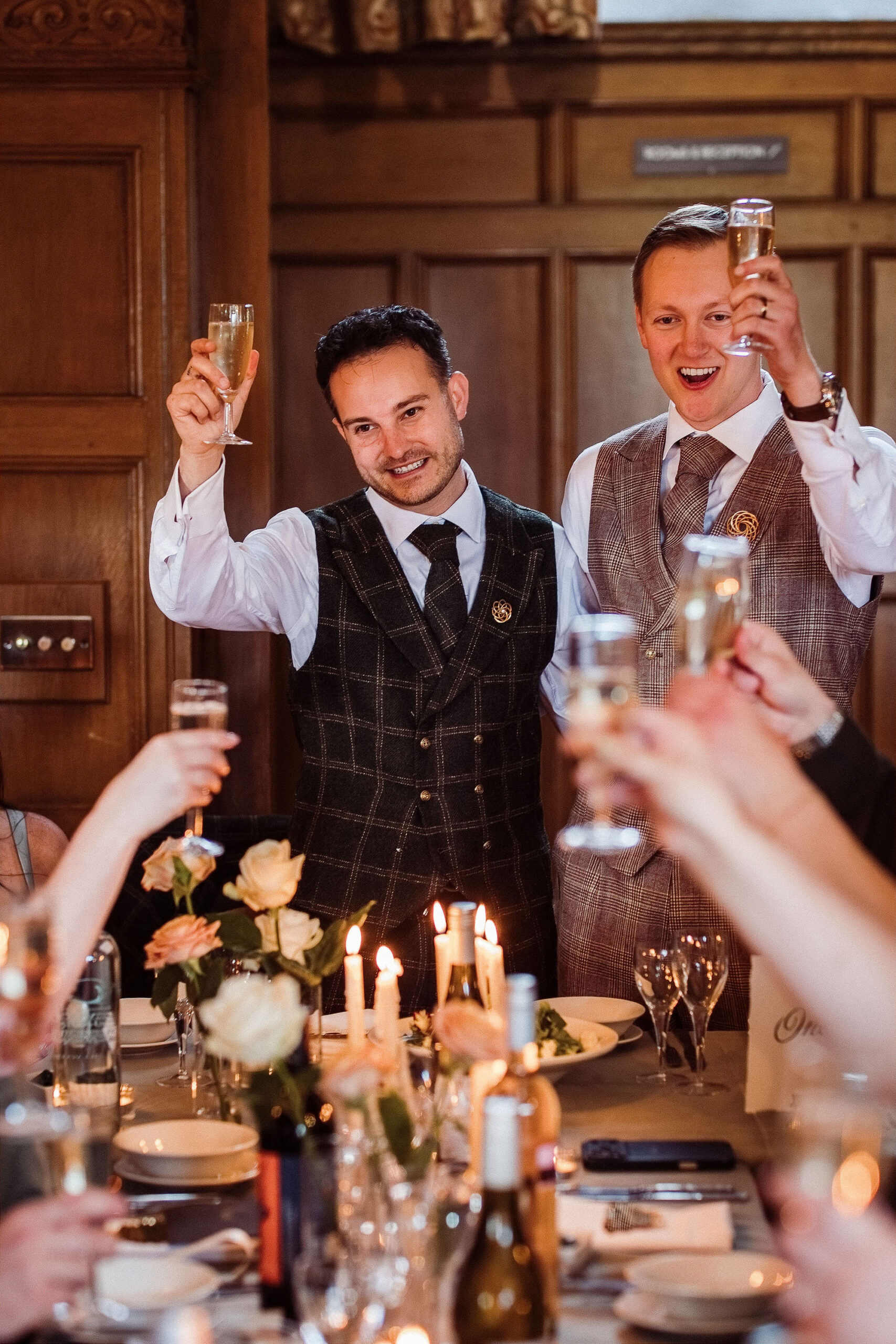 Two grooms smile and raise a toast to their wedding guests at Cragwood Hotel, with roses and candlelight. By Mark Battista Photography