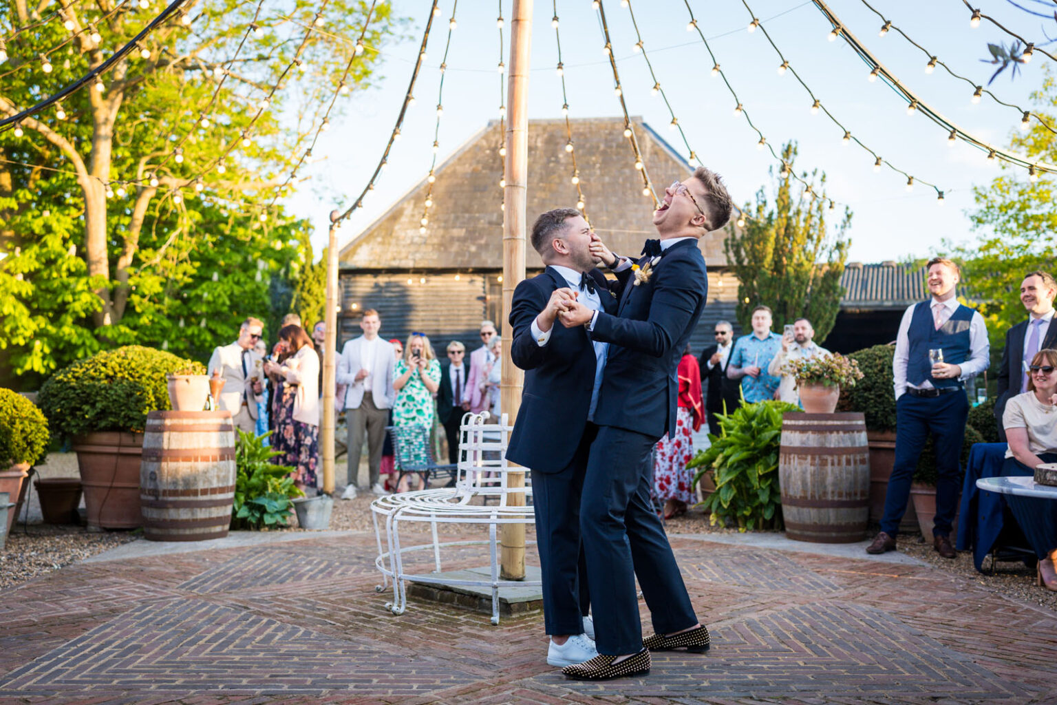 Two grooms laughing as they dance under festoon lights at a Kent wedding venue | Benjamin Toms Photography