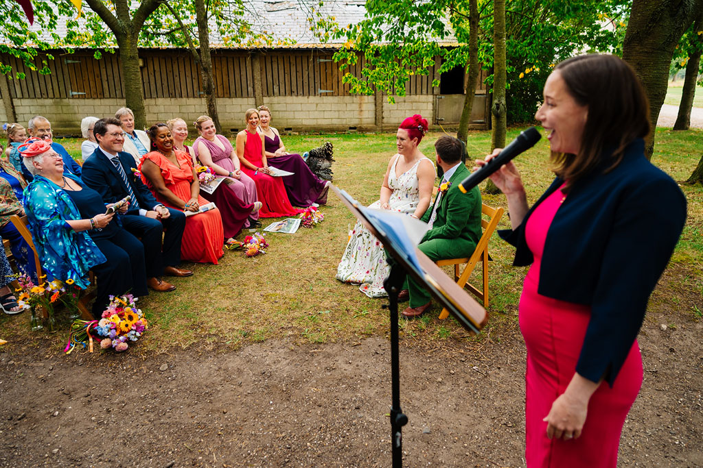 A woman in a red dress speaks into a microphone at an outdoor wedding ceremony in woodland