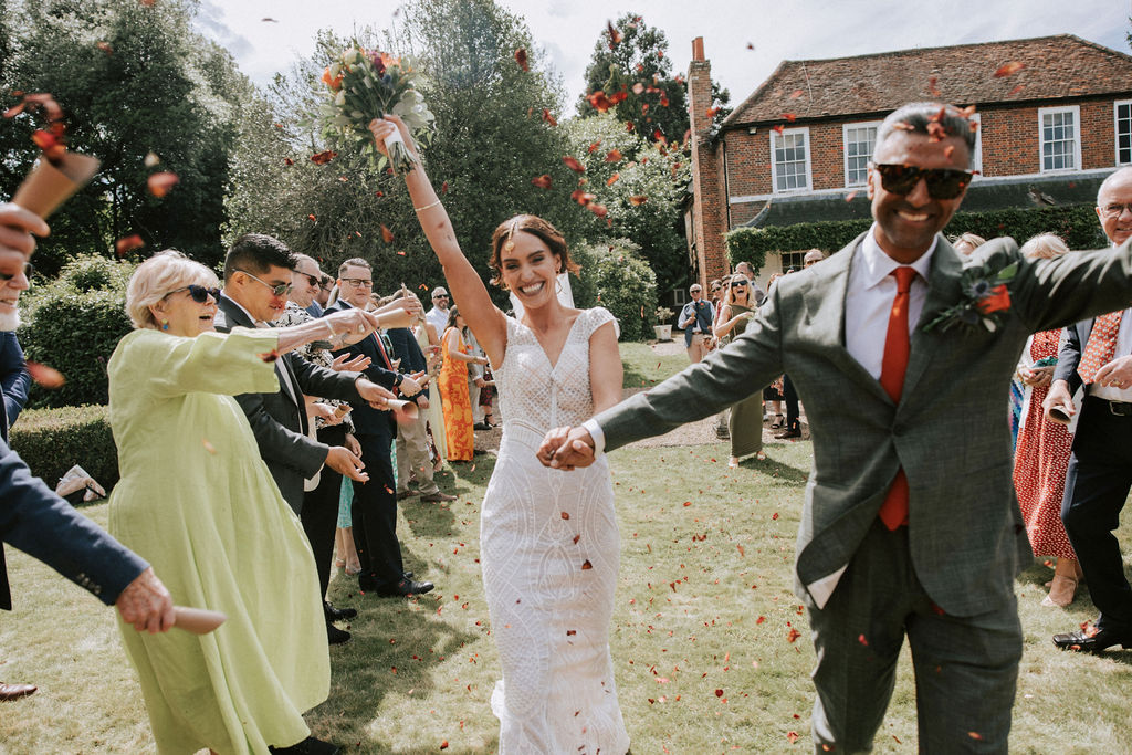 Grinning bride and groom hand in hand on the lawn at Beaumont Hall while guests throw confetti | Nicki Shea Photography