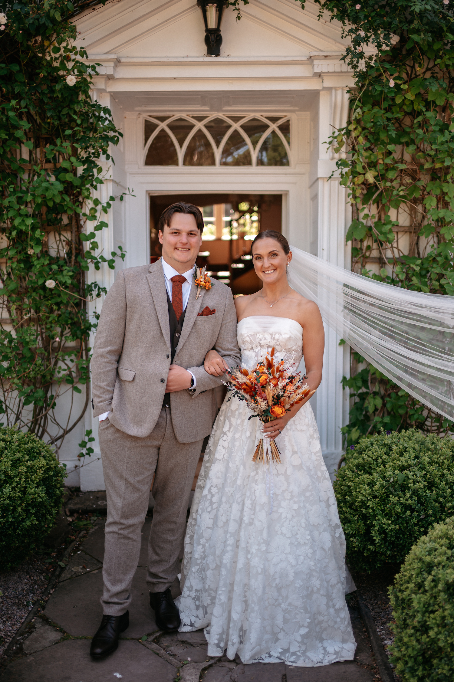 Groom and bride stood by the doorway of their wedding venue. He's wearing a light brown suit with rust coloured tie, she's in a lace dress with a veil and dried flower bouquet | Studio Williams