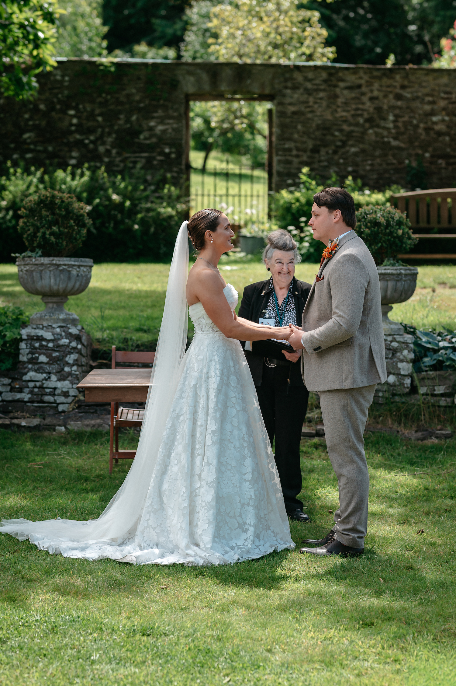 Bride and groom hand in hand on a grassy lawn with wedding registrar smiling as they say their vows | Studio Williams Photography