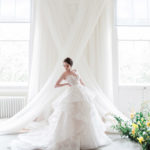 A model in a wedding gown stands before a white wall draped with sheer fabric in a cross between two windows. Credit Amanda Karen Photography