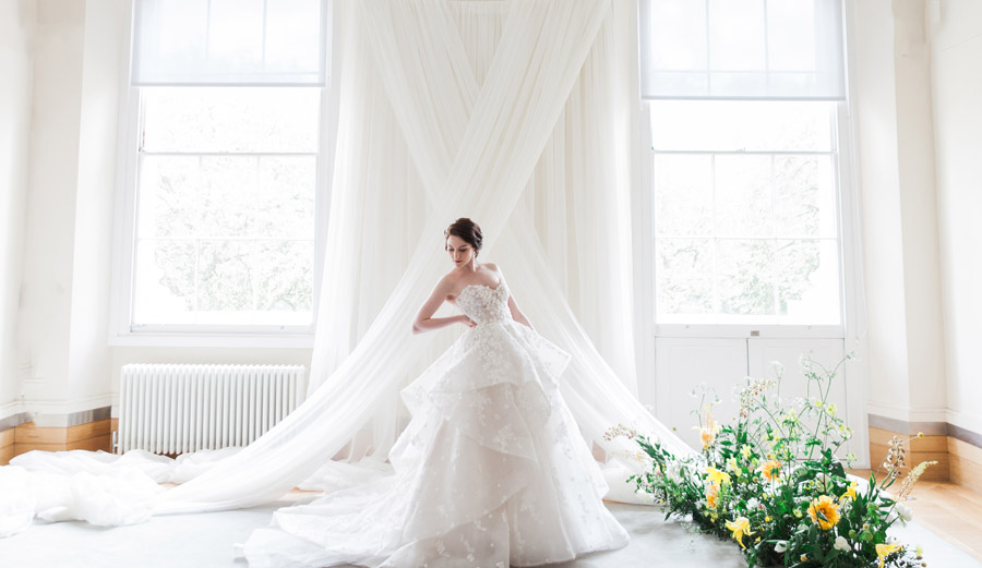 A model in a wedding gown stands before a white wall draped with sheer fabric in a cross between two windows. Credit Amanda Karen Photography