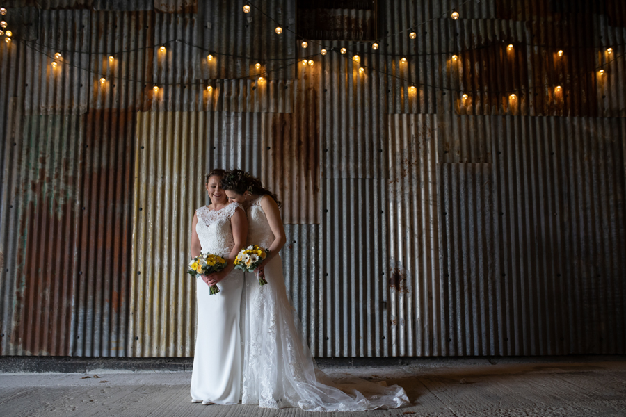 Jess and Hannah wear beautiful complimentary wedding dresses for their day. Both are sleeveless with lace details and long skirts. They look beautiful. They're standing in front of an industrial backdrop with festoon lights. Image by Evolve Photography Devon