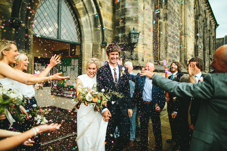 Bride and groom smiling as guests throw confetti outside a church
