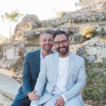 Stephen and Colin after their vow renewal in Greece. Both wear summer suits with shorts, one in a darker blue and one in a pale grey blue. They