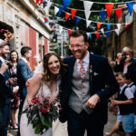 Bride and groom laugh as guests throw confetti. There's red white and blue bunting above