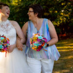 Two brides on their wedding day. The bride on the left wears a white sleeveless dress with a high neck and lace detailing. The bride on the right wears a pastel rainbow sleeveless top in a light fabric, with white trousers. Both hold rainbow coloured posy bouquets and they're beautiful. By Artisan X Photography