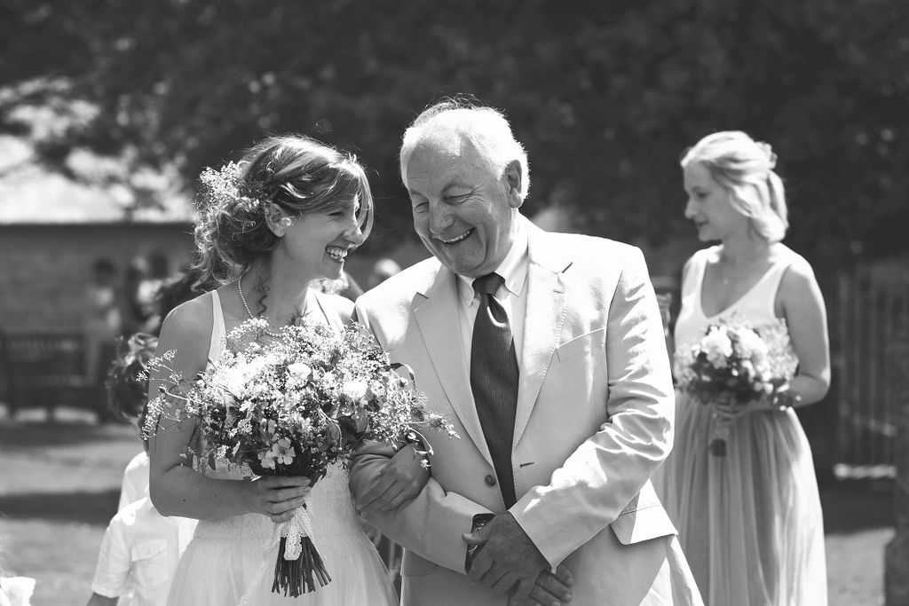 A bride with her dad on her wedding day. They're arm in arm, talking and smiling naturally together. She holds a bouquet of flowers and a bridesmaid walks in the background. Photography credit Lasting Impressions by Lucy