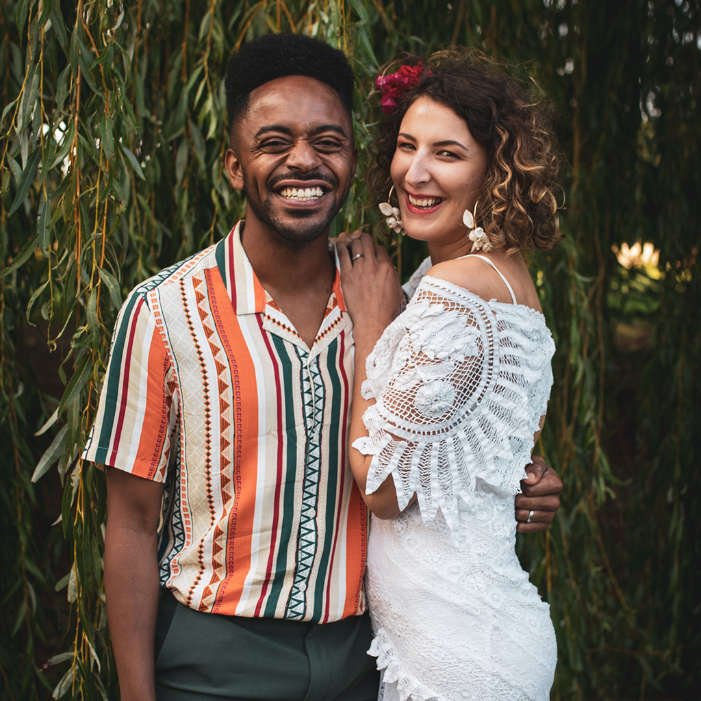 Relaxed wedding couple grinning at the camera, he wears a stripy short sleeved shirt and she's wearing a macrame style boho dress in white