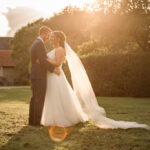 A bride and groom pose for a portrait at golden hour. She's wearing a full length veil which is beautifully lit by the sun. Credit Becky Harley Photography