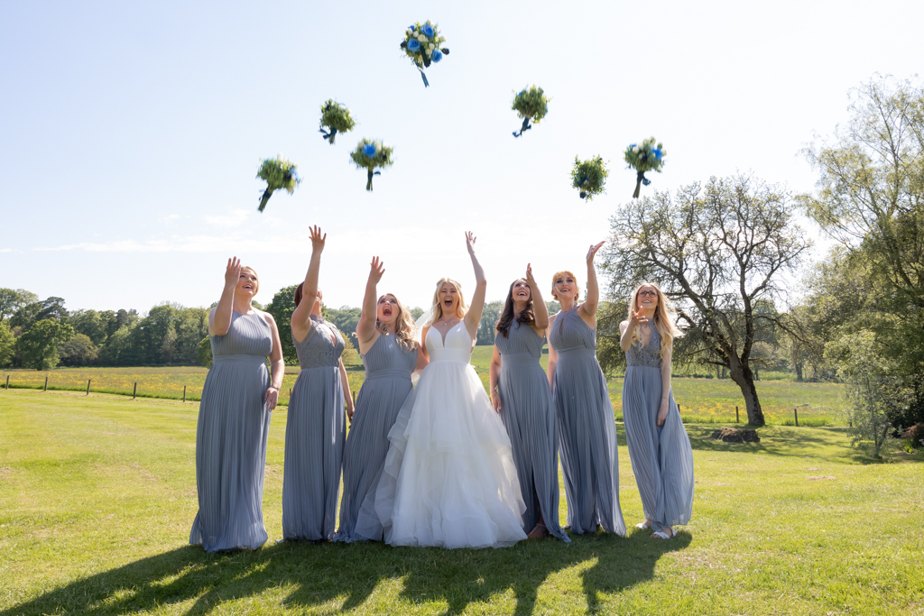 A bride and her six bridesmaids throw their bouquets in the air. The bridesmaids are in pale blue grey dresses. By Evolve wedding photographers in Devon