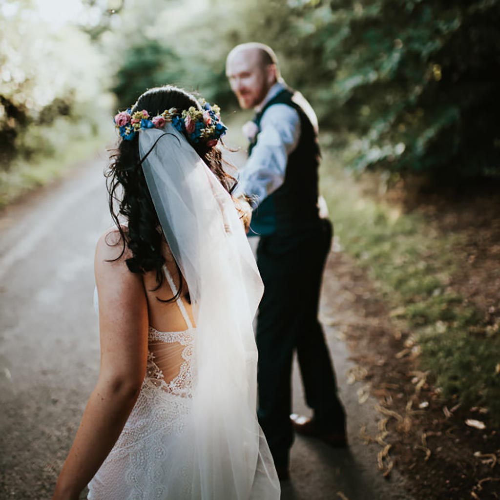 A couple walk along a country road, the sun lights her boho veil and he turns to look back at her. Captured by Beth Beresford Photography