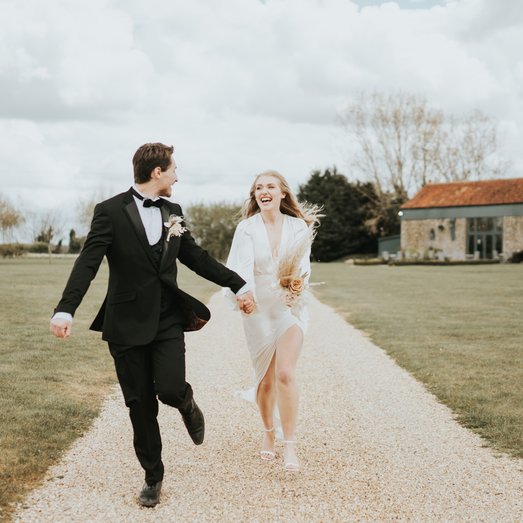 A modern bride and groom run along a path towards the camera, laughing. Captured by Beth Beresford Photography at Pentney Abbey in Norfolk