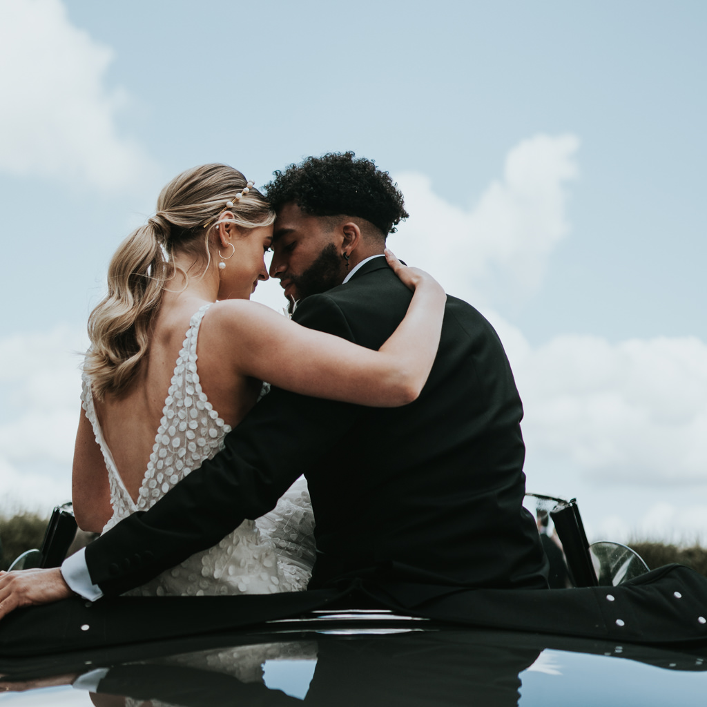 Newlyweds nuzzle noses on the bonnet of the wedding car, their arms around each other. Captured by Beth Beresford Photography at Pentney Abbey in Norfolk