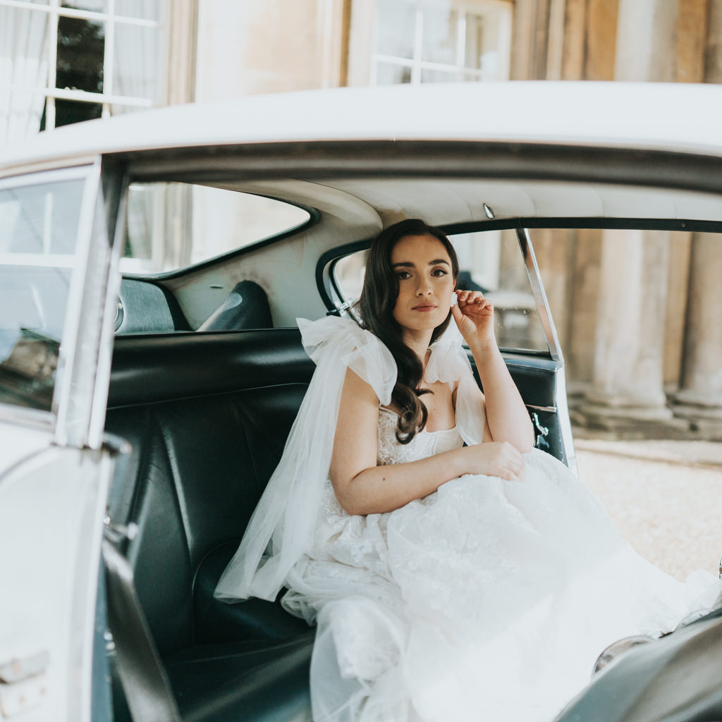 A bride sits in the back of a wedding car, wearing a modern wedding dress and looking thoughtfully towards the camera. Captured by Beth Beresford Photography at The Prestwold Hall in Loughborough