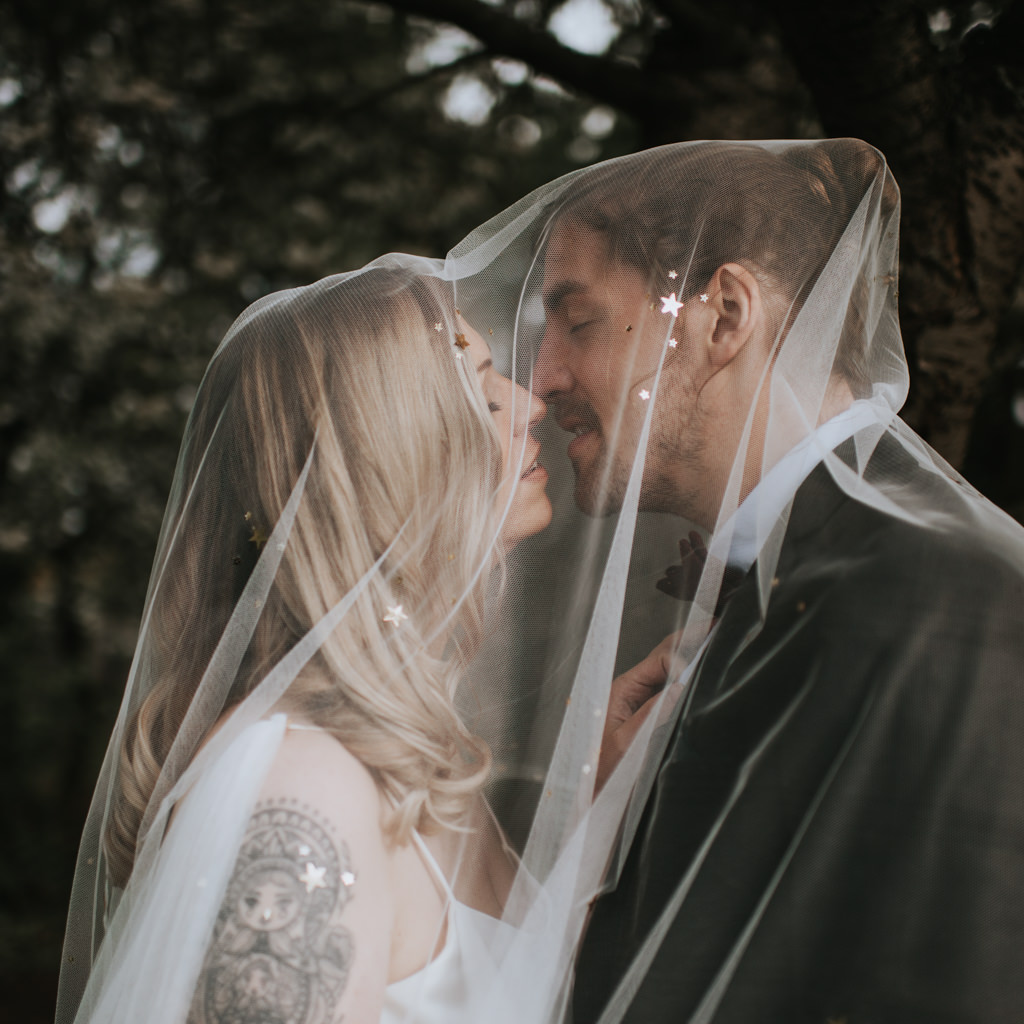 Newlyweds share a kiss under her wedding veil. The bride is blond with a tattoo on her shoulder. Captured by Beth Beresford Photography