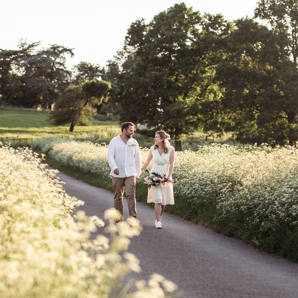 A couple in relaxed wedding outfits walk along a country lane in the evening. Sunlight shines from the cow parsley at the side of the lane, and she carries a pretty bouquet