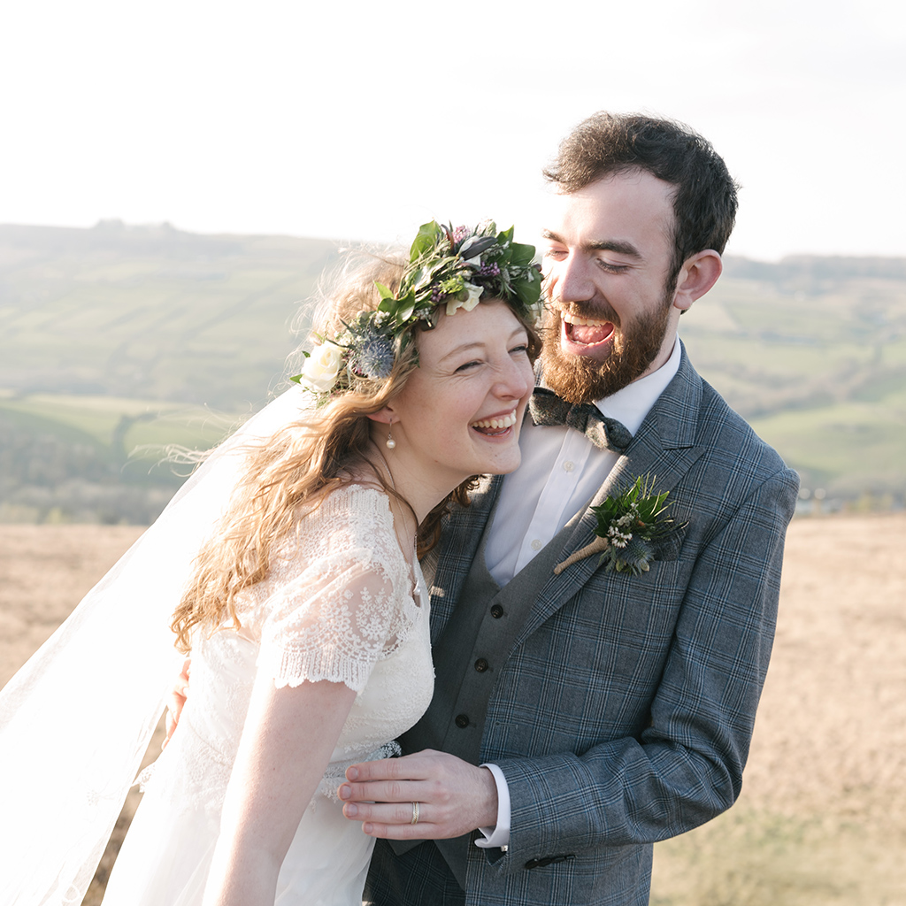 A bride and groom laugh together. They're stood in a corn field, she wears a flower crown and lace dress, he's in a blue grey suit with a bowtie. Credit Hannah Beatrice Photography