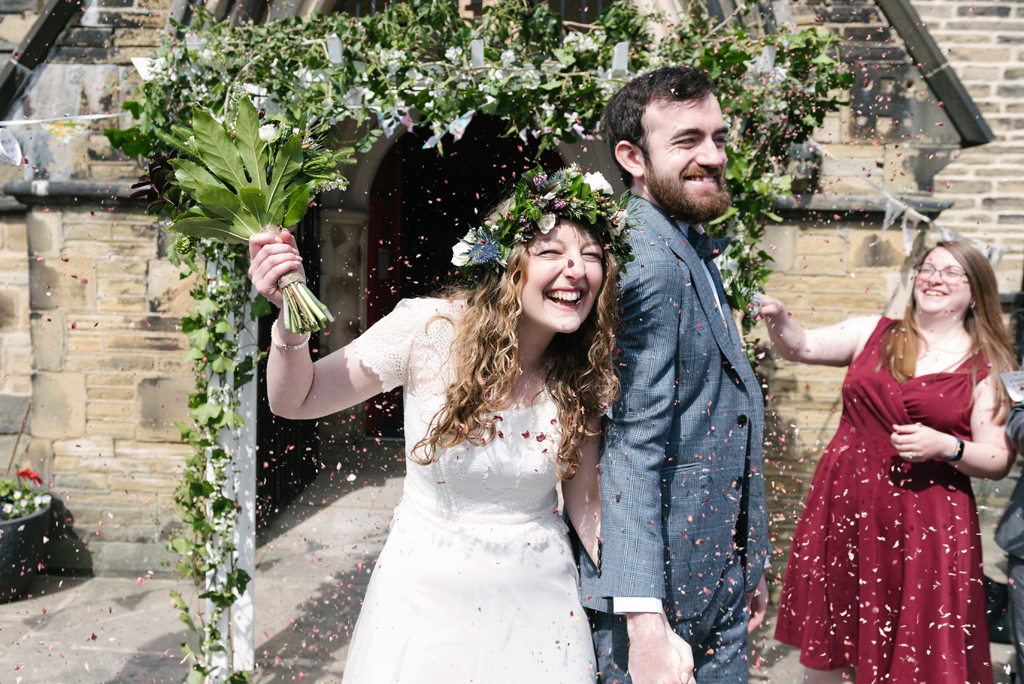 A bride and groom hold hands outside church. Guests are throwing petal confetti, and the bride holds her flowers in the air. They're both grinning; she's beautiful with a floral crown and white dress, while he wears a smart grey suit.