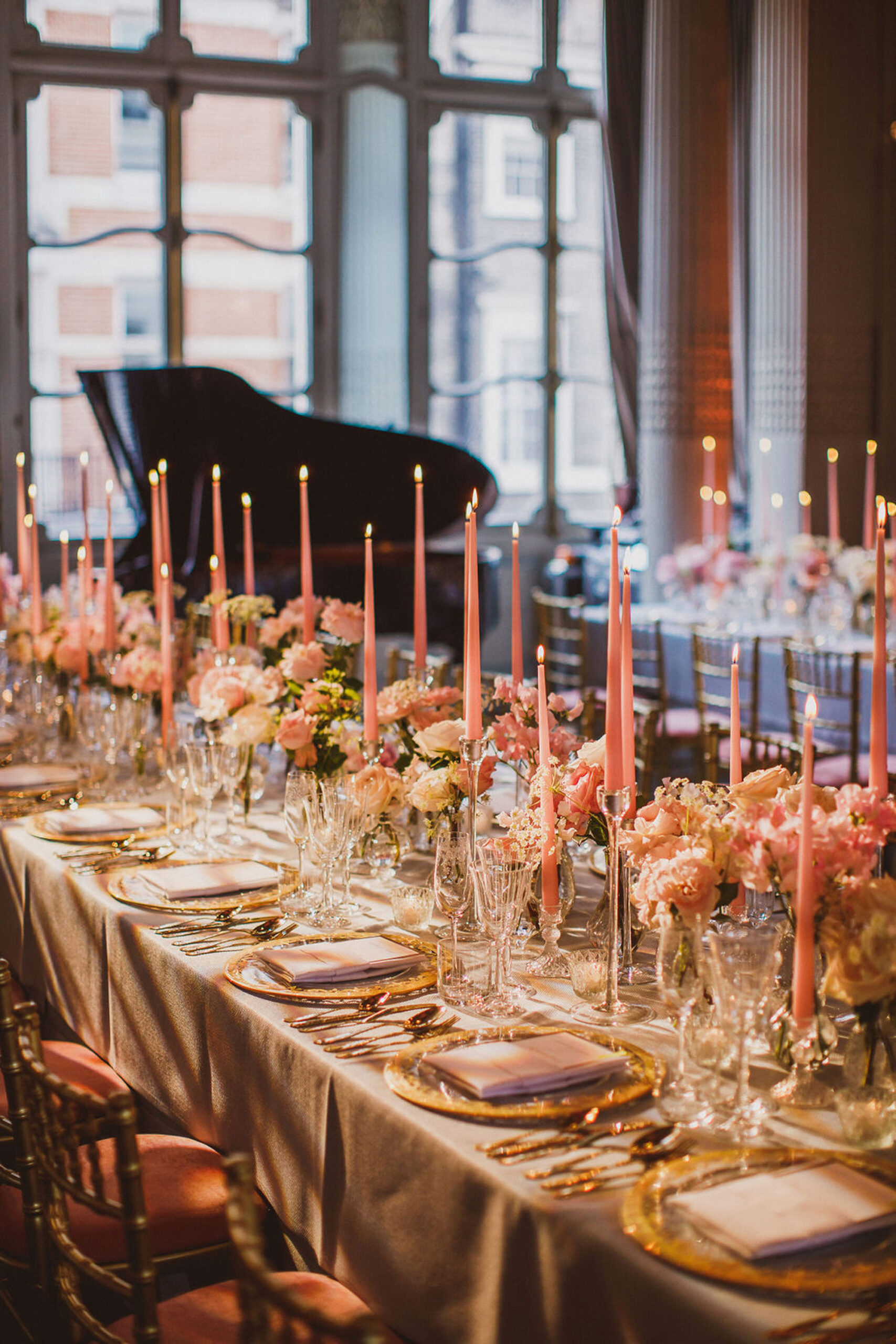 A long banqueting table designed by Mark Niemierko. There are tall pink tapered candles and luxury floral displays running along the tables, and a grand piano in the background